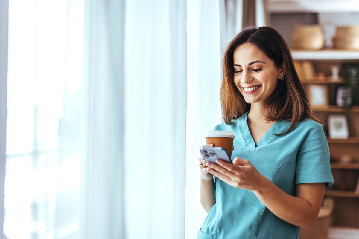 A cheerful nurse in uniform stands near a window, holding a coffee cup and smartphone. She is smiling and appears relaxed, capturing a moment of leisure during a busy day.