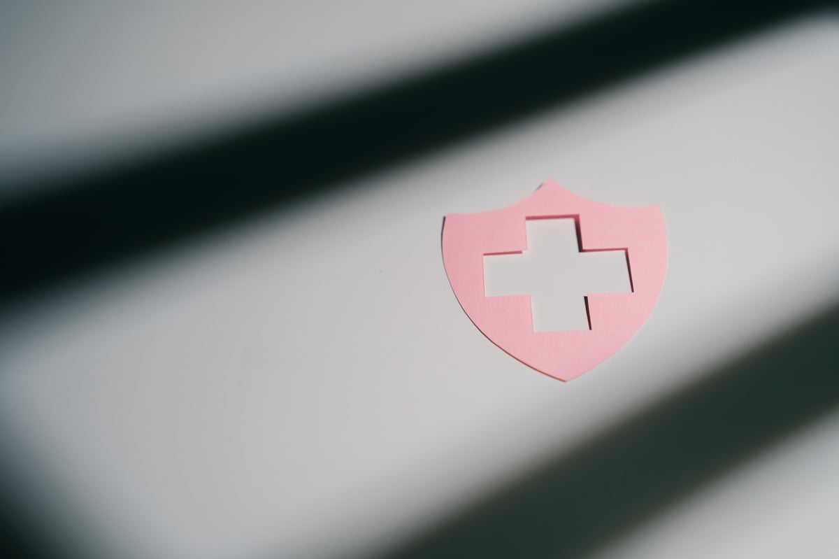 Pink shield with cross cutout on white surface with shadow lines, symbolizing protection and healthcare in calm setting